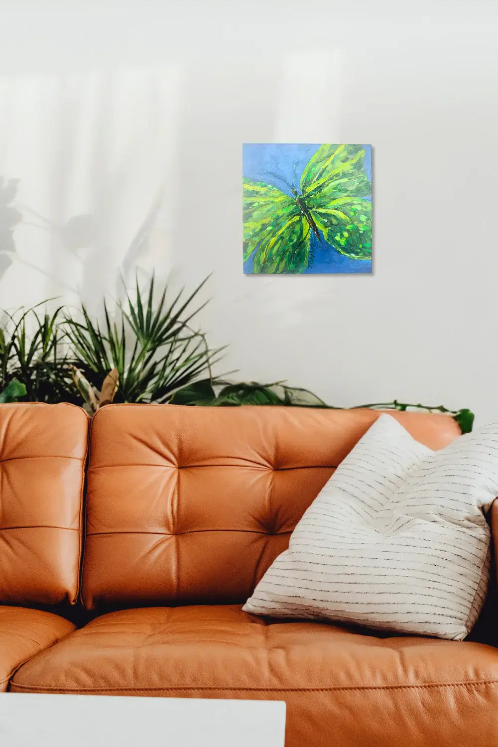 Canvas painting of a green butterfly on a blue background hanging above an orange sofa with a plant in the background. Interior room mock up featuring original painting "Emerge" by Teri Stephens