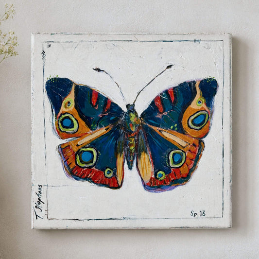 A square canvas painting of a butterfly with dark blue, orange, red, and green patterned wings on a white textured background, mounted on a light beige wall. Original butterfly painting by artist Teri Stephens titled Buckeye