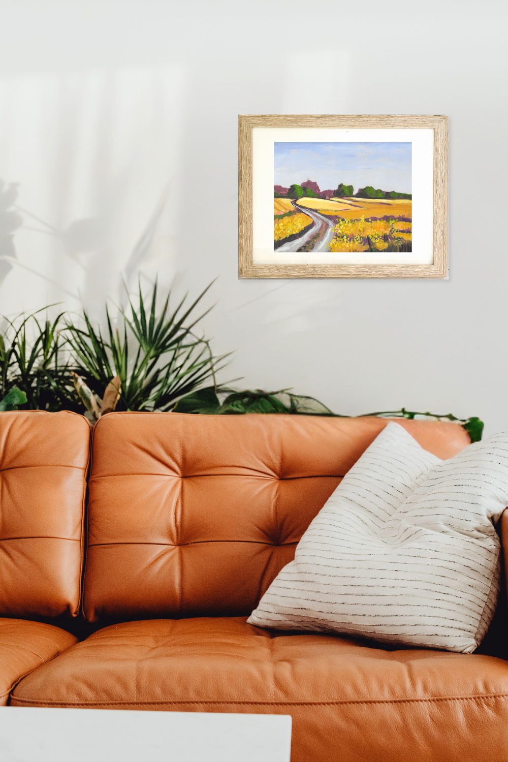 Framed painting of a field with a path on a wall above an orange sofa. Original rural landscape titled Canola by Teri Stephens