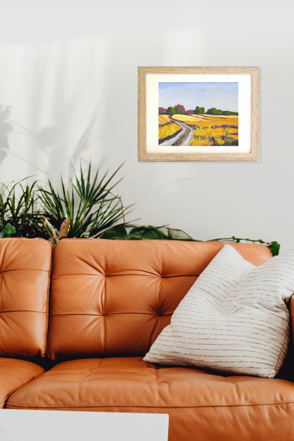Framed painting of a field with a path on a wall above an orange sofa. Original rural landscape titled Canola by Teri Stephens