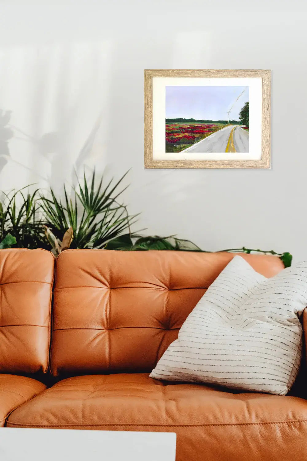 Framed artwork of a road through a field on a wall above an orange sofa with a plant in the corner.