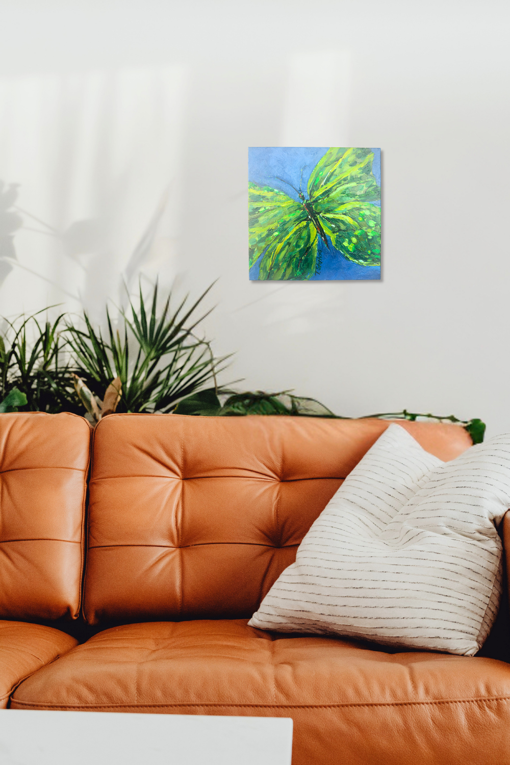 Canvas painting of a green butterfly on a blue background hanging above an orange sofa with a plant in the background. Interior room mock up featuring original painting "Emerge" by Teri Stephens