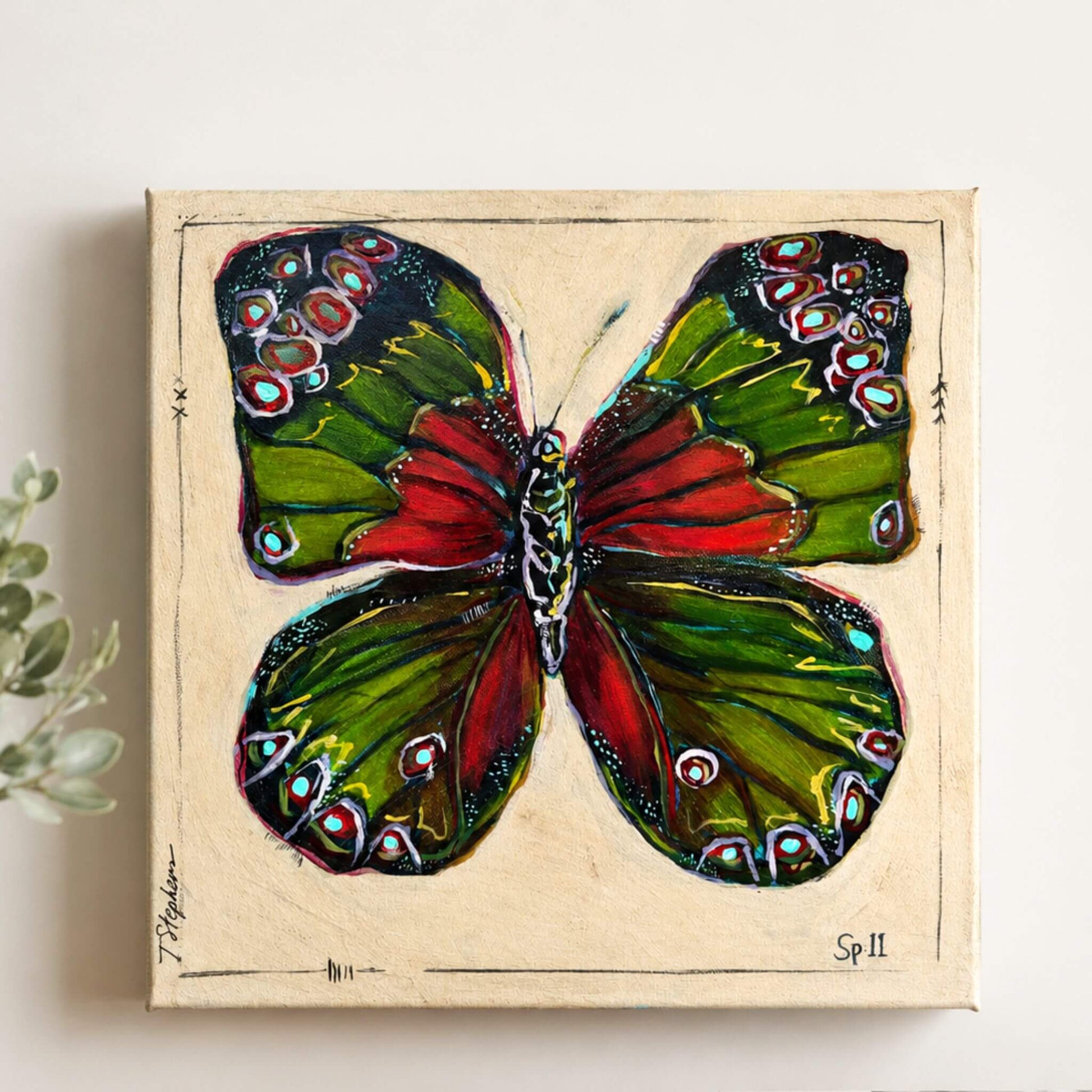A square canvas painting of a butterfly with green and red wings, decorated with colorful spots, mounted on a light beige wall next to a small green plant.