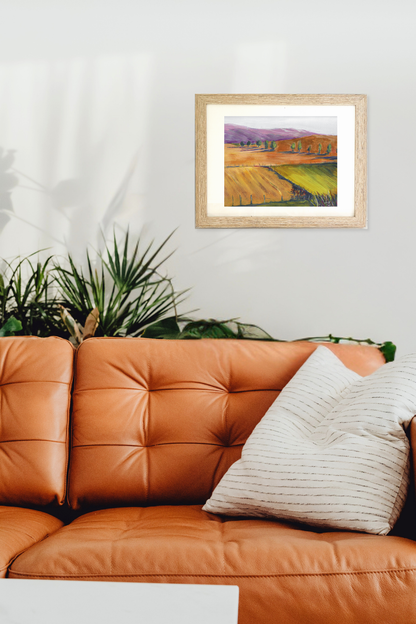Framed landscape painting above an orange leather sofa with a plant in the background "Fallow" by Teri Stephens