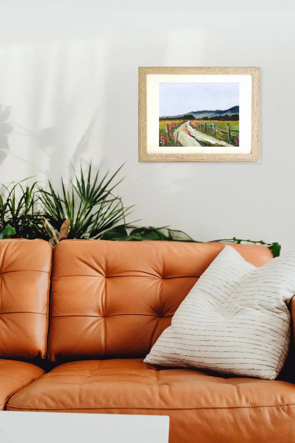Framed landscape painting above an orange leather sofa with a plant in the background.