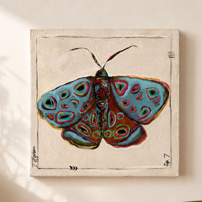 A square canvas painting of a colorful painted moth with blue, green, red, and orange details on a textured cream background, hanging on a beige wall.