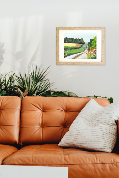 Framed artwork of a garden scene above an orange leather sofa with decorative pillows. "Lane" by artist Teri Stephens