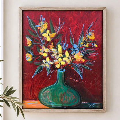 A framed painting of a green vase filled with yellow and blue flowers against a textured red background, hung on a light beige wall with a small plant leaf visible at the lower left. Titled "Maybe" by artist Teri Stephens