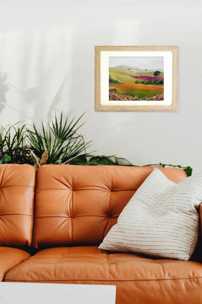 Framed artwork of a landscape above an orange leather sofa with a plant in the corner.