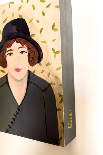Decorative wall art with a stylized portrait of a woman wearing a black hat on a leaf-patterned background. Rue by artist Teri Stephens