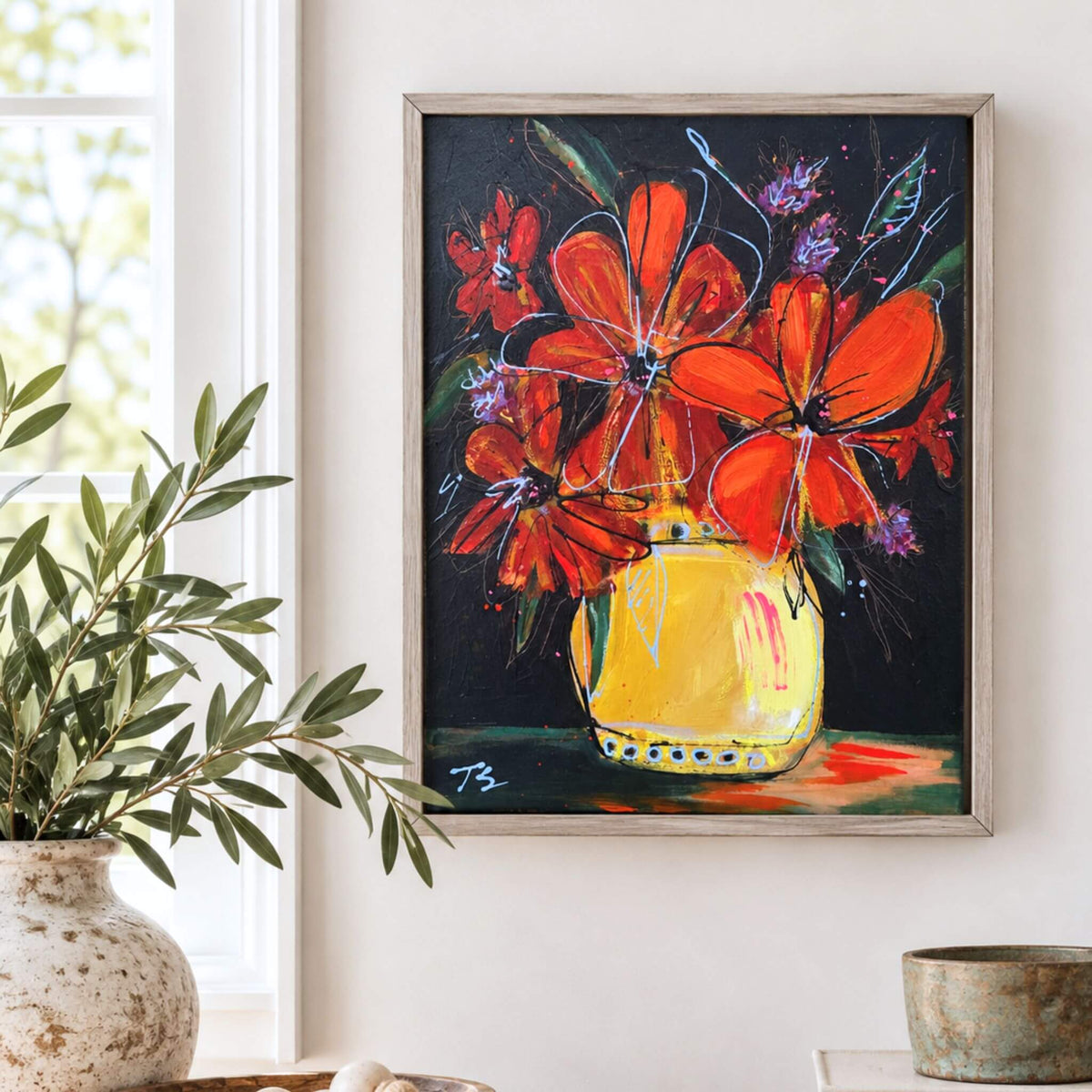 A framed floral painting hanging on a white wall, showing bright orange-red flowers in a yellow vase against a dark background. A potted green plant and a rustic bowl sit nearby in a styled interior setting.