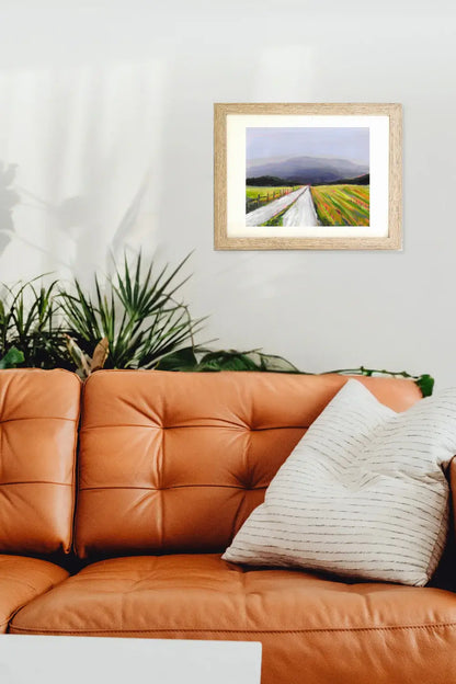 Framed artwork of a landscape with a road and mountains above an orange sofa with a plant and pillow.